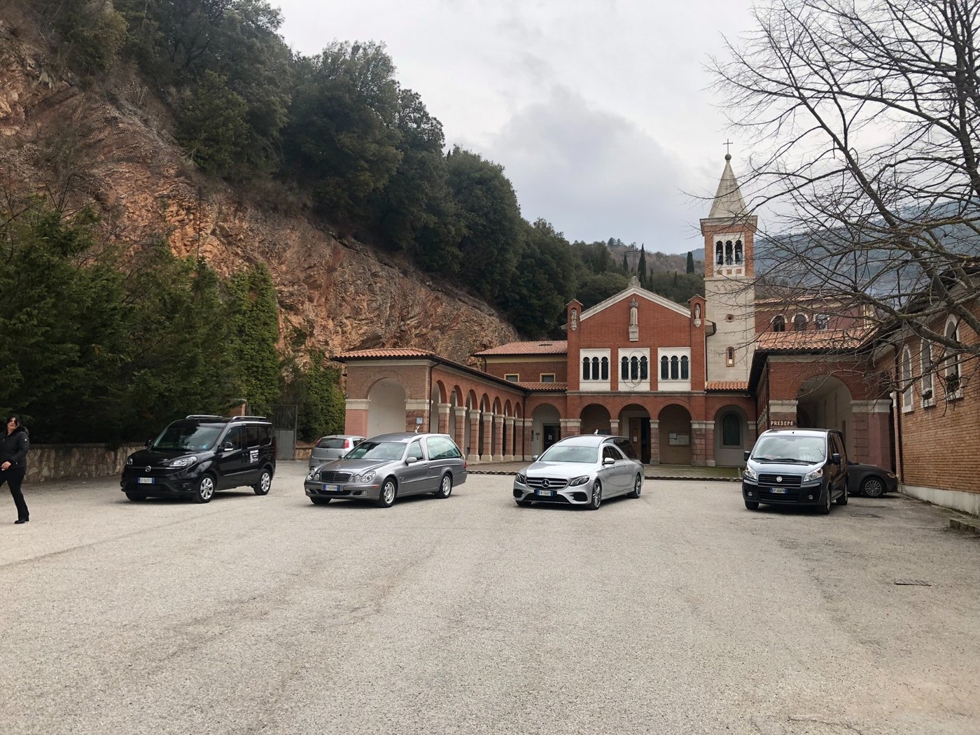 Due Mercedes grigio argento e due furgoni neri davanti a una chiesa in mattoni rossi con portico ad archi e campanile