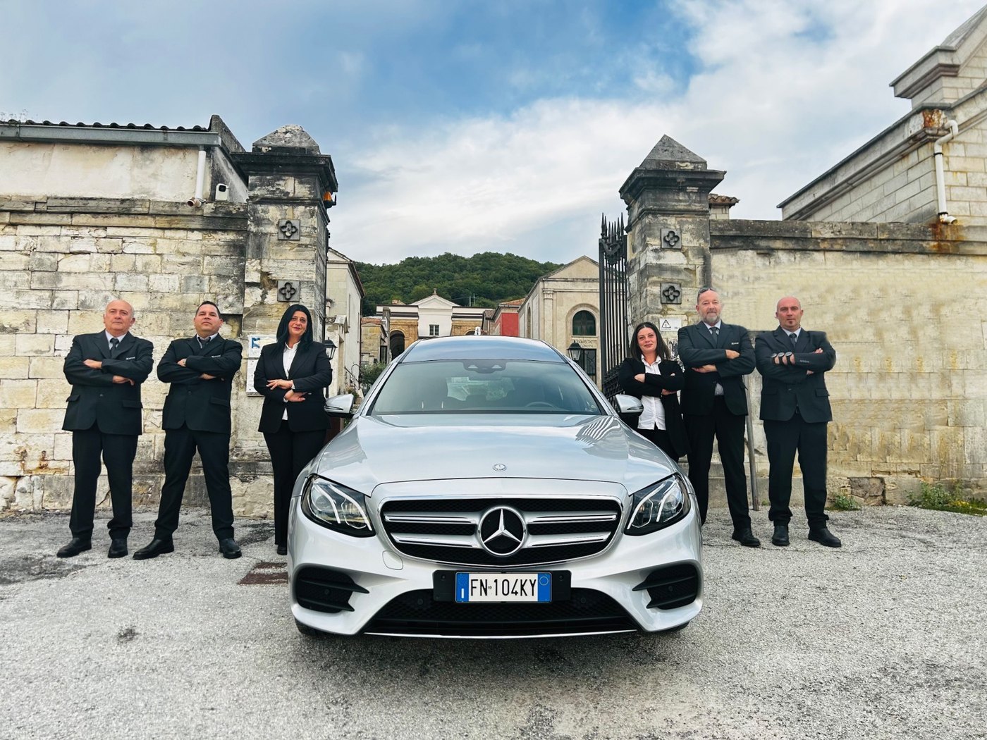 Sei addetti Morroni in abito scuro attorno a una Mercedes argento targata FN104KY, davanti a un portale in pietra